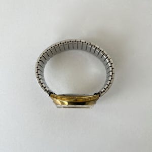 May include: A wristwatch featuring a circular gold-tone metal case and a flexible silver-tone stainless steel expansion band. The watch is positioned to show the side profile of the case and the connected links of the metallic stretch strap.