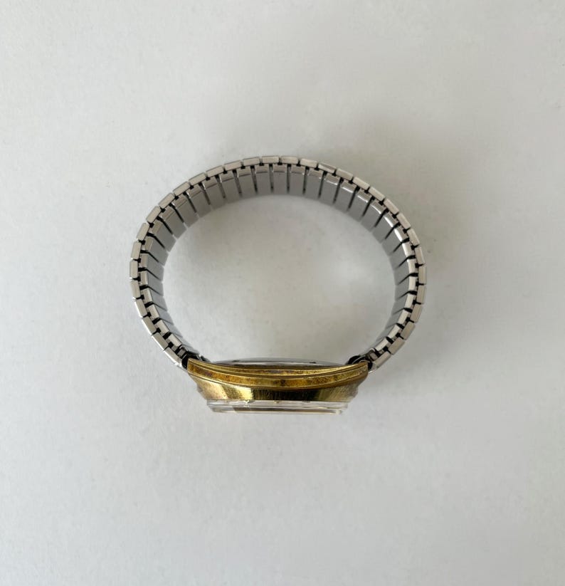 May include: A wristwatch featuring a circular gold-tone metal case and a flexible silver-tone stainless steel expansion band. The watch is positioned to show the side profile of the case and the connected links of the metallic stretch strap.