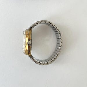 May include: A wristwatch featuring a gold-tone metal case and a silver-tone expandable metal link bracelet. The side view shows the circular watch face with a clear crystal cover and a small winding crown positioned on the right side of the case.