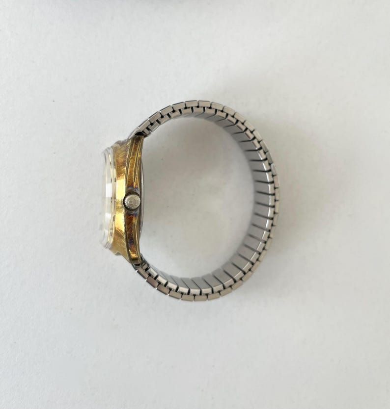 May include: A wristwatch featuring a gold-tone metal case and a silver-tone expandable metal link bracelet. The side view shows the circular watch face with a clear crystal cover and a small winding crown positioned on the right side of the case.
