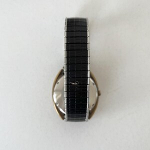 May include: A wristwatch featuring a gold-tone case and a black textured expansion band. The watch has a metallic silver-colored back casing and a flexible, segmented strap with a repeating rectangular pattern across the entire length.
