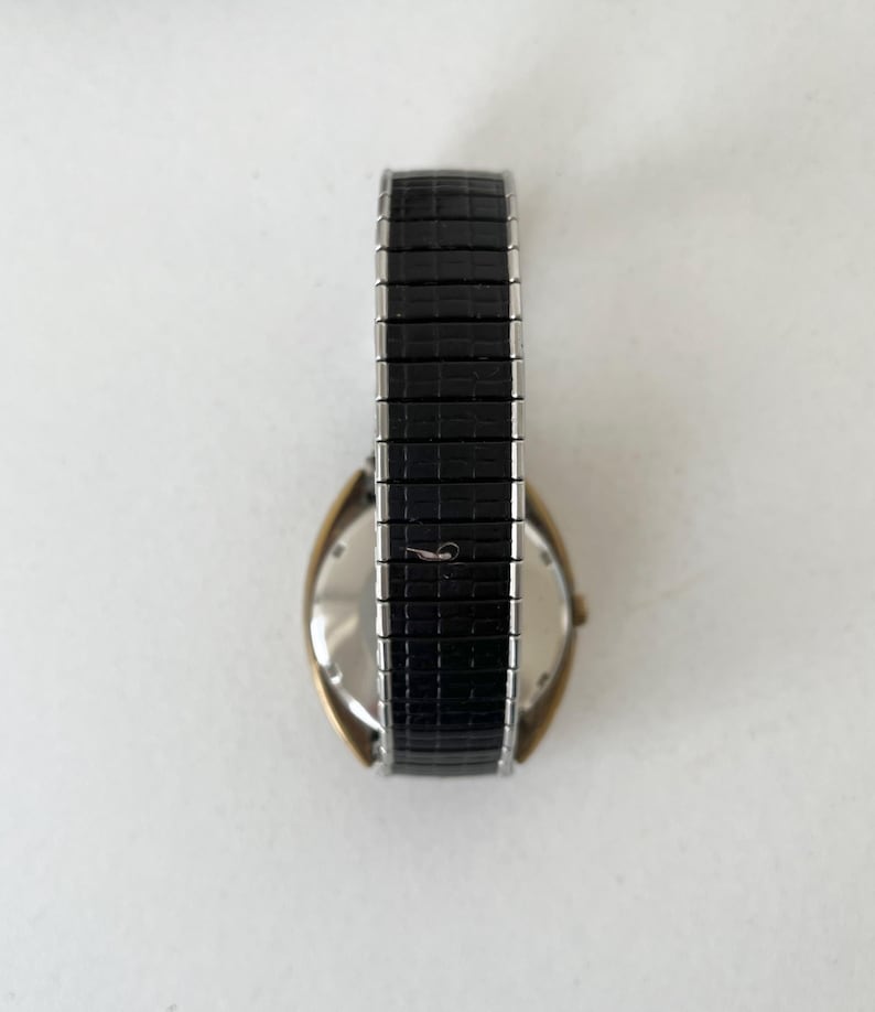 May include: A wristwatch featuring a gold-tone case and a black textured expansion band. The watch has a metallic silver-colored back casing and a flexible, segmented strap with a repeating rectangular pattern across the entire length.