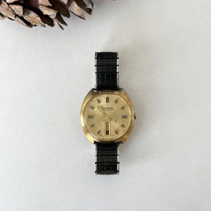 May include: A gold-tone analog wristwatch with a round face, a sunburst champagne dial, and black hour markers. The watch features a day-date window at the six o'clock position and is attached to a black, textured, expandable metal link strap.