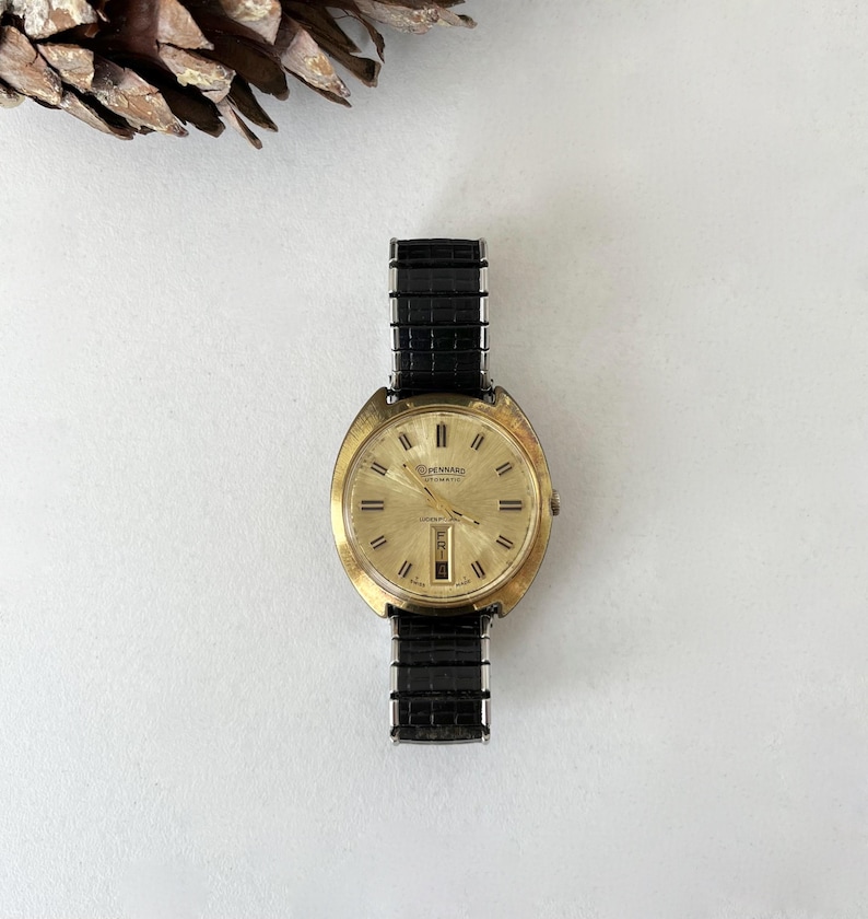 May include: A gold-tone analog wristwatch with a round face, a sunburst champagne dial, and black hour markers. The watch features a day-date window at the six o'clock position and is attached to a black, textured, expandable metal link strap.
