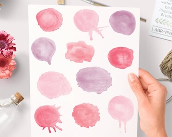 May include: A sheet of paper with 12 watercolor blots in shades of pink and purple. The blots are arranged in a grid pattern. The paper is surrounded by various craft supplies, including a cup of coffee, a spool of twine, and a paintbrush.