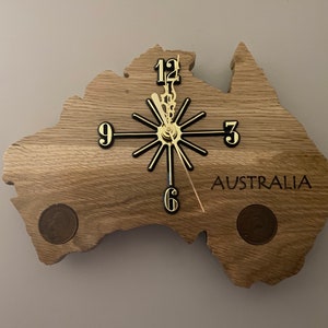 May include: Wooden wall clock shaped like the map of Australia. The clock has a gold-colored clock face and black hands. The word "AUSTRALIA" is printed on the clock face. Two coins are embedded in the wood.