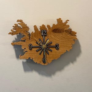 May include: A wooden wall clock in the shape of Iceland. The clock has black numerals at the 12, 3, 6, and 9 o'clock positions, with a starburst design at the centre. The clock hands are black and gold.