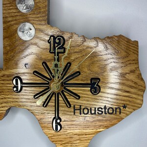 May include: A wooden clock shaped like the state of Texas with gold-colored hands and black numbers. The clock face has a black starburst design. The clock is inscribed with the word "Houston*" in black lettering.