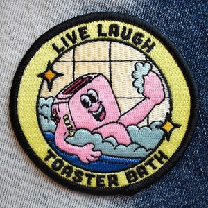 May include: A yellow and black embroidered patch with a cartoon toaster in a bathtub. The text on the patch reads "Live Laugh Toaster Bath".