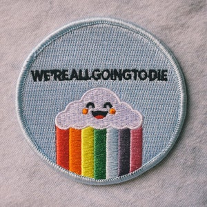 Könnte beinhalten: Ein blauer gestickter Aufnäher mit einer Regenbogenwolke und dem Text "WE'RE ALL GOING TO DIE".