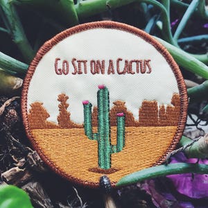 May include: A round, white embroidered patch with a brown border. The patch features a green cactus with pink flowers and the text "Go Sit On A Cactus".