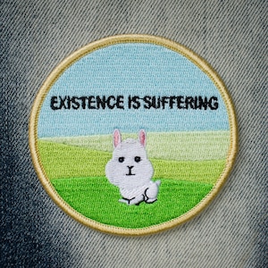 Puede incluir: Un parche redondo bordado con un conejito blanco en un campo verde. El parche tiene un fondo de cielo azul y el texto "EXISTENCE IS SUFFERING" en negro.