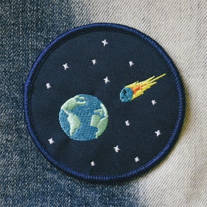 May include: A blue embroidered patch with a white outline depicting a blue and green Earth with a yellow and orange comet flying past it on a dark blue background with white stars.