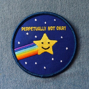 May include: A blue and yellow embroidered patch with a smiling yellow star shooting across a rainbow. The text "PERPETUALLY NOT OKAY" is embroidered in white on the patch.
