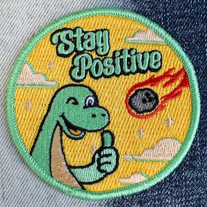 May include: Green embroidered patch with a yellow background. The patch features a cartoon dinosaur with a thumbs up and the text "Stay Positive".