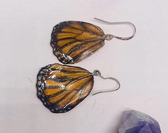May include: A pair of earrings featuring real monarch butterfly wings, set in silver with a simple hook closure. The wings are a vibrant orange with black veins and white spots.