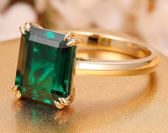 Op de afbeelding: Een gouden ring met een grote, smaragdgeslepen, diepgroene edelsteen. De ring heeft een eenvoudig ontwerp met een gouden band en klauwen die de edelsteen vasthouden. De ring staat tegen een zachte, vage achtergrond.