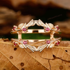 Könnte beinhalten: Ein Ringverstärker aus Roségold mit einem zarten Blattdesign und kleinen roten Edelsteinen. Der Ringverstärker ist so konzipiert, dass er um einen größeren Verlobungsring passt.