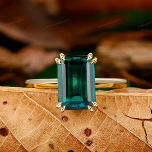 Può includere: Un anello d'oro con una grande pietra preziosa verde smeraldo tagliata a smeraldo. L'anello è appoggiato su una foglia marrone.