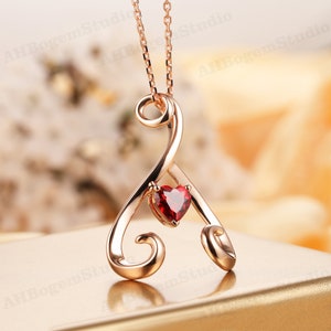 Puede incluir: Un collar de oro rosa con un delicado colgante en forma de corazón adornado con una piedra preciosa roja. El colgante está diseñado con un patrón de espiral intrincado.
