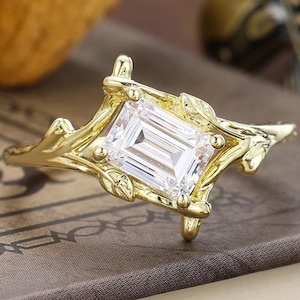 Puede incluir: Un anillo dorado con una gema clara de talla esmeralda. El anillo presenta un diseño inspirado en la naturaleza, con hojas y ramas. El anillo se encuentra sobre un fondo neutro.