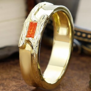 Puede incluir: Un anillo dorado con una piedra naranja rectangular en el centro. El anillo tiene un diseño texturizado y orgánico alrededor de la piedra. El anillo está sobre una superficie marrón, con un libro y una miniatura de la Torre Eiffel en el fondo.