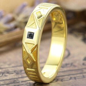 May include: A gold-colored ring with geometric patterns and a small, square, black gemstone. The ring has a polished finish and is set against a blurred background with hints of purple and brown.