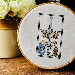 May include: A white embroidery hoop with a cross-stitch design of a sword, crown, and flowers. The design is in a rectangular frame with a white background and colorful details.