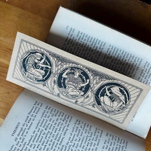 May include: A brown bookmark featuring a black and white illustration of three figures in circles. The figures are surrounded by vines and leaves. The text "THESEUS" is at the top of the bookmark.