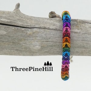 May include: A rainbow colored metal chainmail bracelet with a silver clasp. The bracelet is displayed on a piece of driftwood. The text "ThreePineHill" is visible below the driftwood.