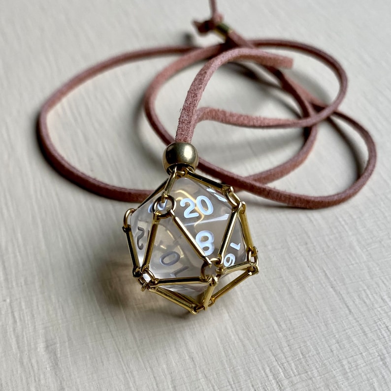 May include: A clear glass twenty-sided die with gold metal framing, hanging from a brown leather cord. The die has white numbers on each side.
