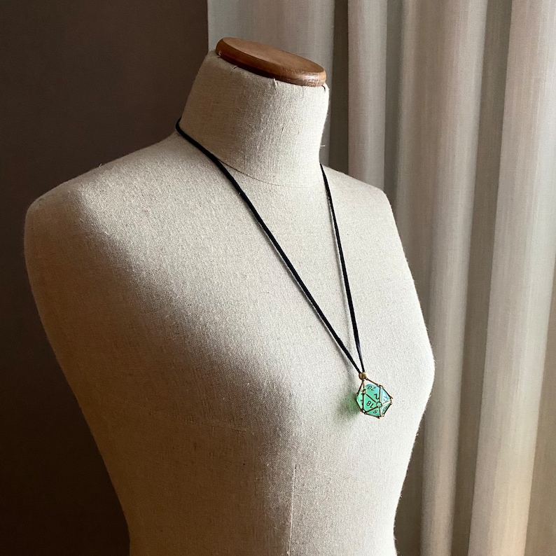 May include: A black cord necklace with a green glass pendant shaped like a dice. The pendant has gold wire accents.