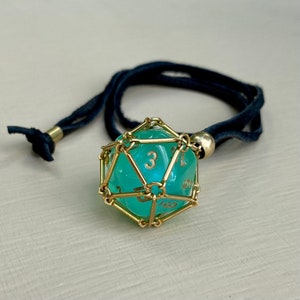 May include: A turquoise d20 dice pendant with gold wire cage, hanging from a black cord necklace.
