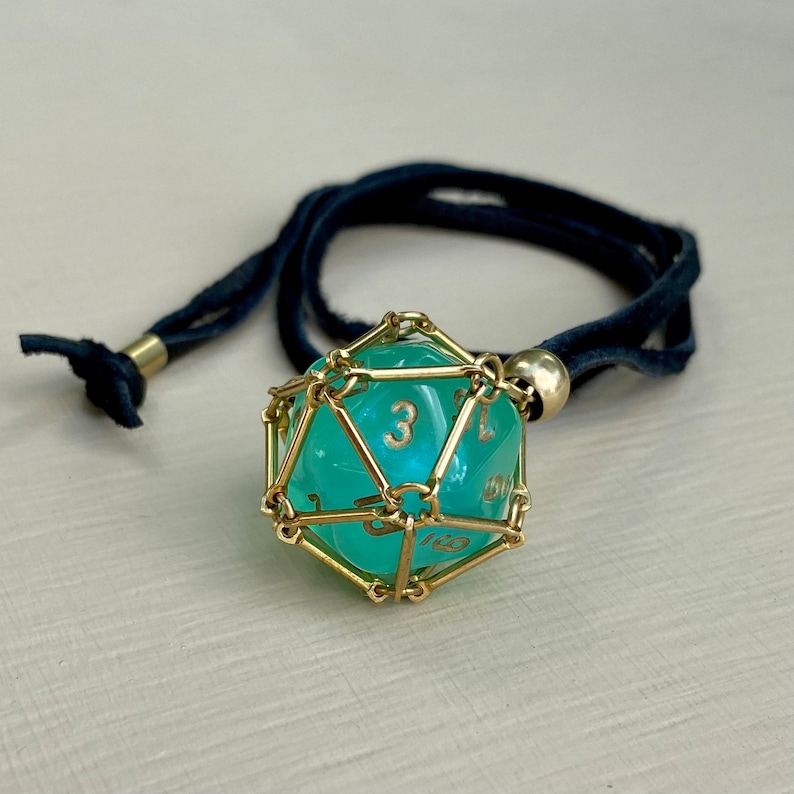 May include: A turquoise d20 dice pendant with gold wire cage, hanging from a black cord necklace.