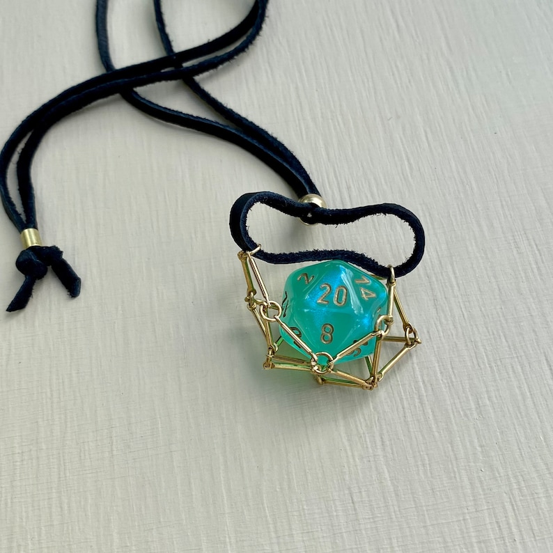 May include: A turquoise 20-sided die with gold numbers is suspended in a gold wire cage. The cage is attached to a black leather cord necklace.