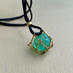 May include: A green, translucent dice with gold numbers is encased in a gold wire cage. The dice is hanging from a black leather cord.