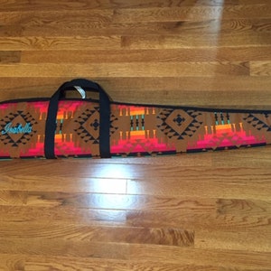 May include: A brown, black, red, orange, and turquoise patterned gun case with a black handle and the name "Isabella" embroidered on the front.