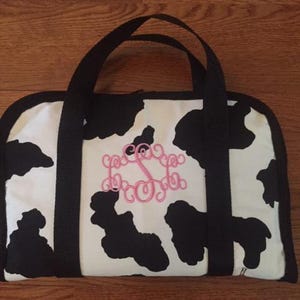 May include: A white and black cow-print tote bag with black handles and trim. The bag features a pink monogram in the center. The bag is rectangular with rounded corners.