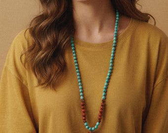 May include: A long turquoise and red beaded necklace with a green tassel. The necklace is worn over a mustard yellow t-shirt. The person is smiling and wearing a maroon headband. The background is a solid tan color.