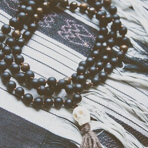 A black mala necklace with a skull pendant and a tassel, made of lava, bone, yak wool, and bronzite. The necklace is displayed on a black and white striped textile. The text "black kali" is at the bottom.