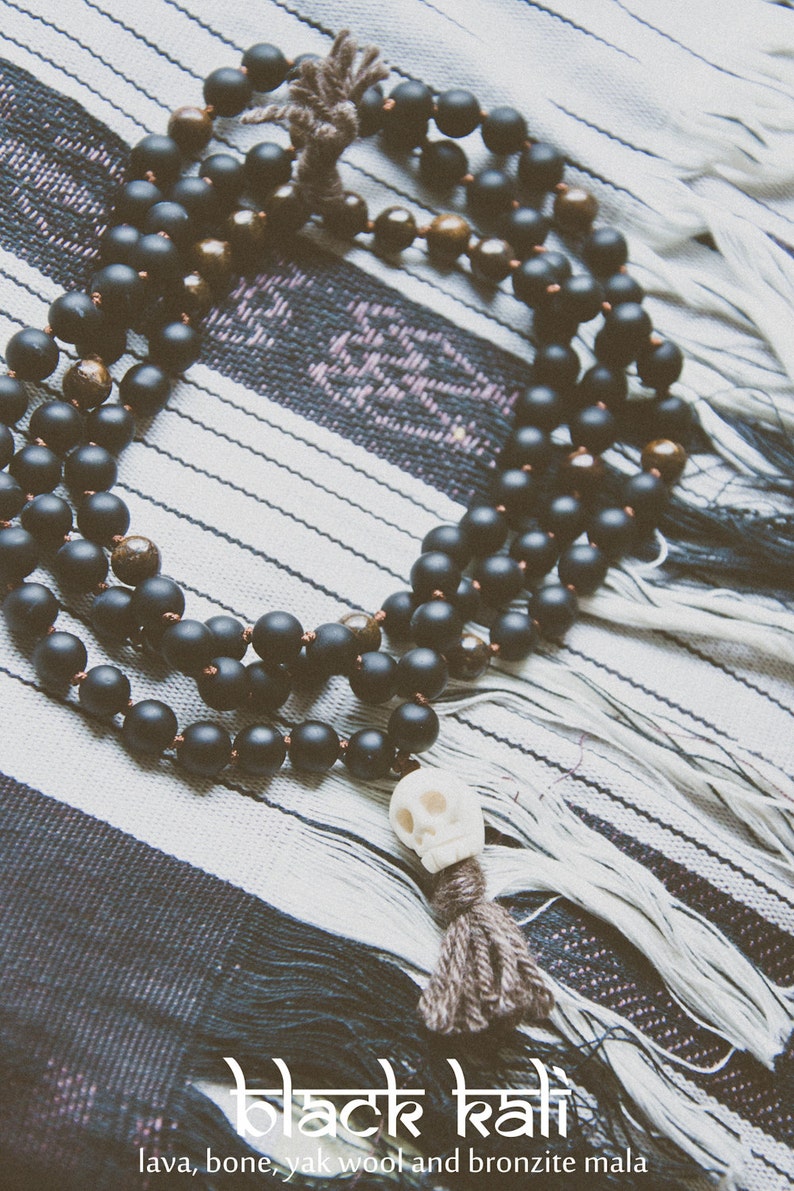 A black mala necklace with a skull pendant and a tassel, made of lava, bone, yak wool, and bronzite. The necklace is displayed on a black and white striped textile. The text "black kali" is at the bottom.
