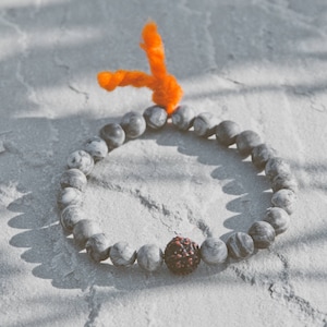 Könnte beinhalten: Ein Armband mit grauen und weißen Steinperlen und einer einzelnen dunkelroten Perle. Das Armband ist mit einem orangefarbenen Faden gebunden. Der Text "NOMAD ACCESSORIES" ist im Bild zu sehen.