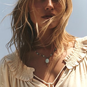 May include: A woman wearing a cream-colored blouse with a ruffled neckline and a delicate floral pattern. She is also wearing a necklace with a large, oval, light blue pendant. Her blonde hair is flowing in the wind.