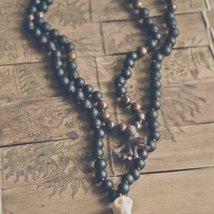 A black beaded necklace with a skull pendant and tassel. The necklace features matte black beads and brown beads, with a skull-shaped pendant and a gray tassel. The necklace is displayed on a wooden surface. Black Kali Mala Necklace worn by a man • Lava & Bronzite 108 Beads Yak Wool Meditation beads • Carved Bone Skull Jewelry
