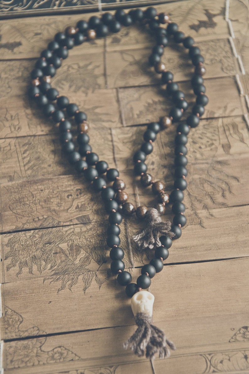 A black beaded necklace with a skull pendant and tassel. The necklace features matte black beads and brown beads, with a skull-shaped pendant and a gray tassel. The necklace is displayed on a wooden surface. Black Kali Mala Necklace worn by a man • Lava & Bronzite 108 Beads Yak Wool Meditation beads • Carved Bone Skull Jewelry