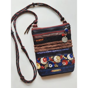 Op de afbeelding: Een rechthoekige crossbodytas met een zwarte rits, bruine en zwarte leren details en bloemenborduurwerk. Het heeft een rood-zwarte touwriem en een bedel met een munt en een blauwe steen. De tekst "NOMAD MINI CROSSBODY" staat onderaan.
