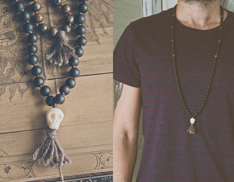 A long beaded necklace with black matte beads, a white skull pendant, and a gray tassel. The necklace is displayed on a wooden surface and worn by a person.