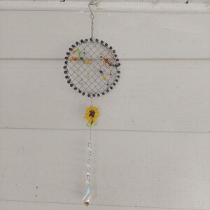 May include: A handmade dreamcatcher with a circular wire frame, adorned with black beads, colorful accents, and a yellow sunflower charm. A chain of clear beads and a prism dangle below. The dreamcatcher hangs from a metal hook.