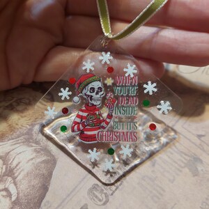 May include: Clear, diamond-shaped Christmas ornament with a skeleton wearing a Santa hat and striped sweater. The ornament has the text "When You're Dead Inside But It's Christmas" and snowflake and dot accents. A green and white ribbon is attached.