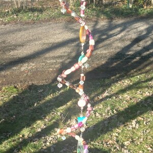 May include: A handcrafted wind chime with a spiral design. It features a variety of colorful beads in shades of pink, white, brown, and blue, with a large, oval-shaped pendant at the bottom. The chime is suspended outdoors.